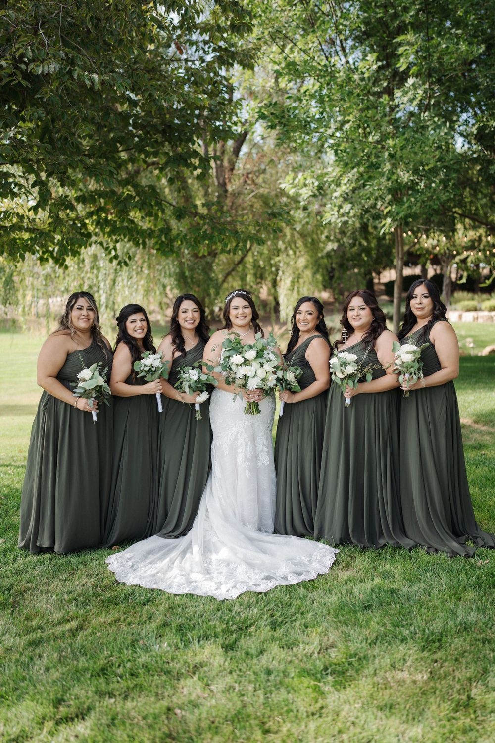 Bridal party in sage green dresses posing outdoors with bride in white lace gown.