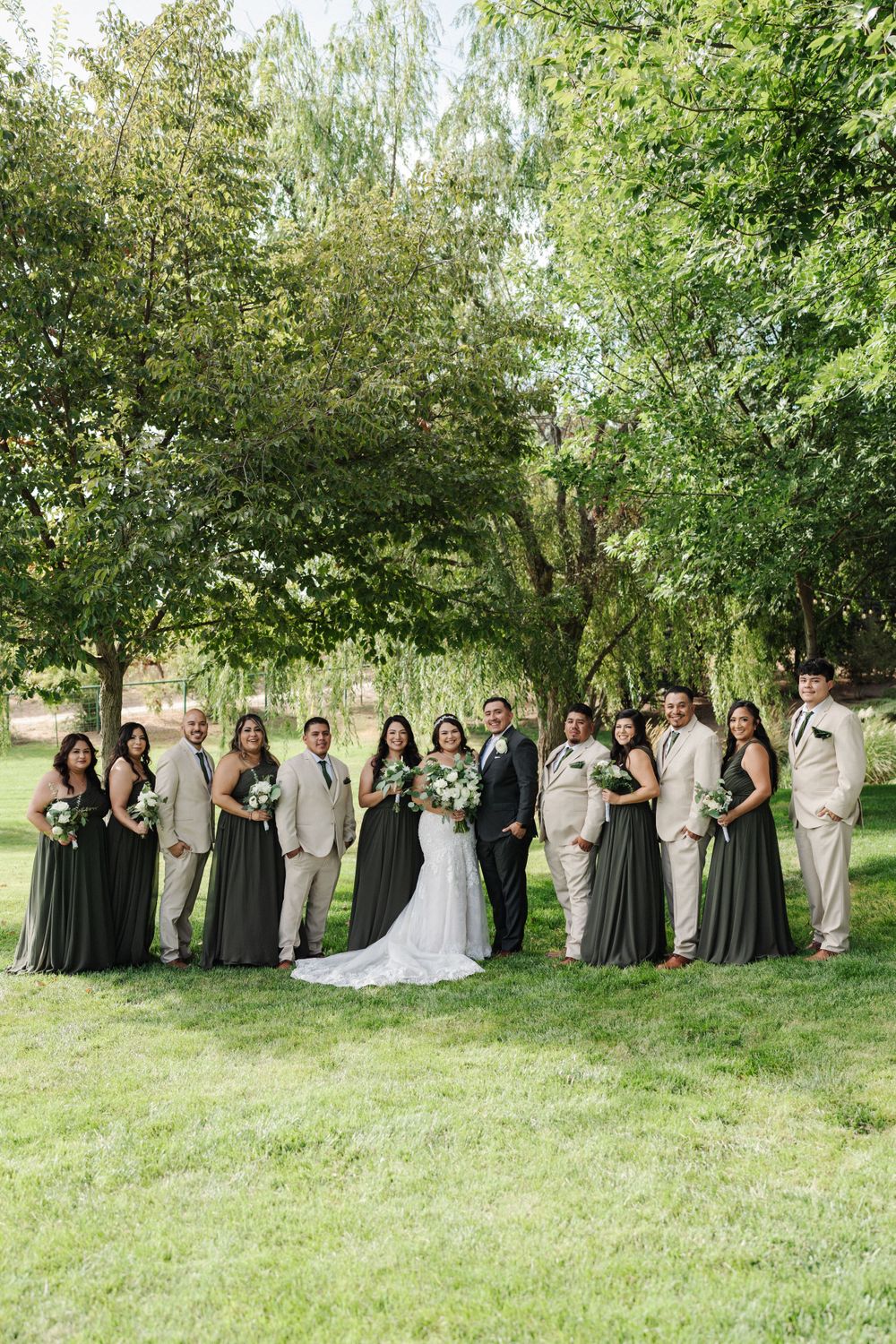 Wedding party in green dresses and tan suits posing together on grassy lawn.