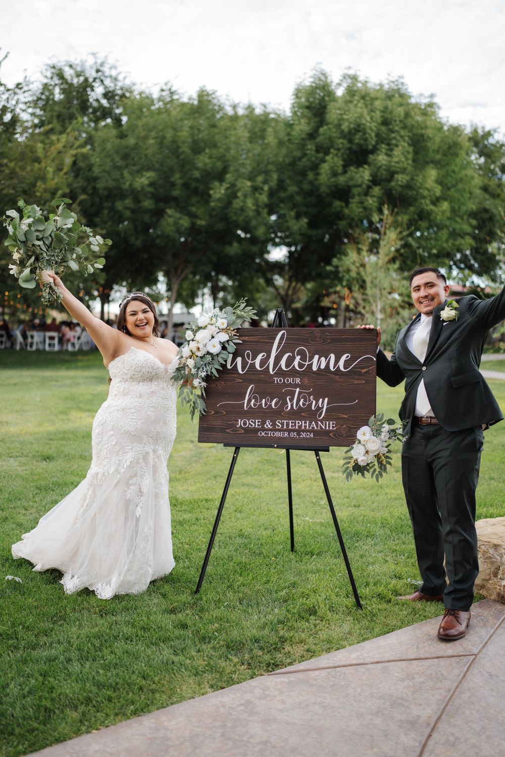 Newlyweds celebrating by welcome sign with floral decorations at outdoor venue.