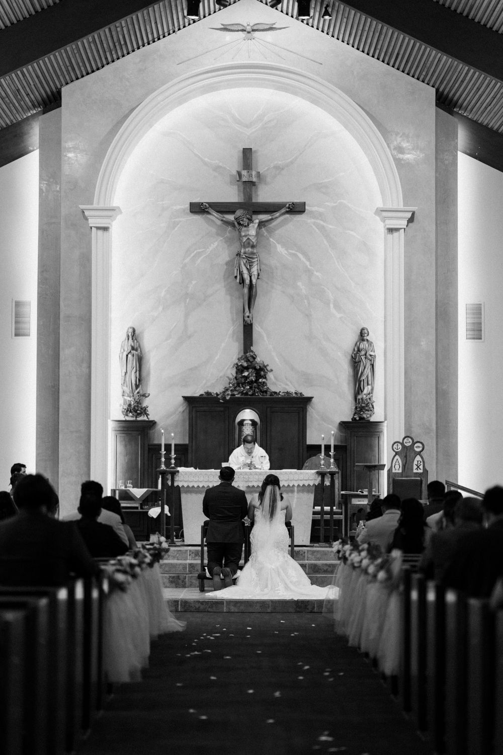 Black and white photo of wedding ceremony in traditional church with ornate crucifix altar.