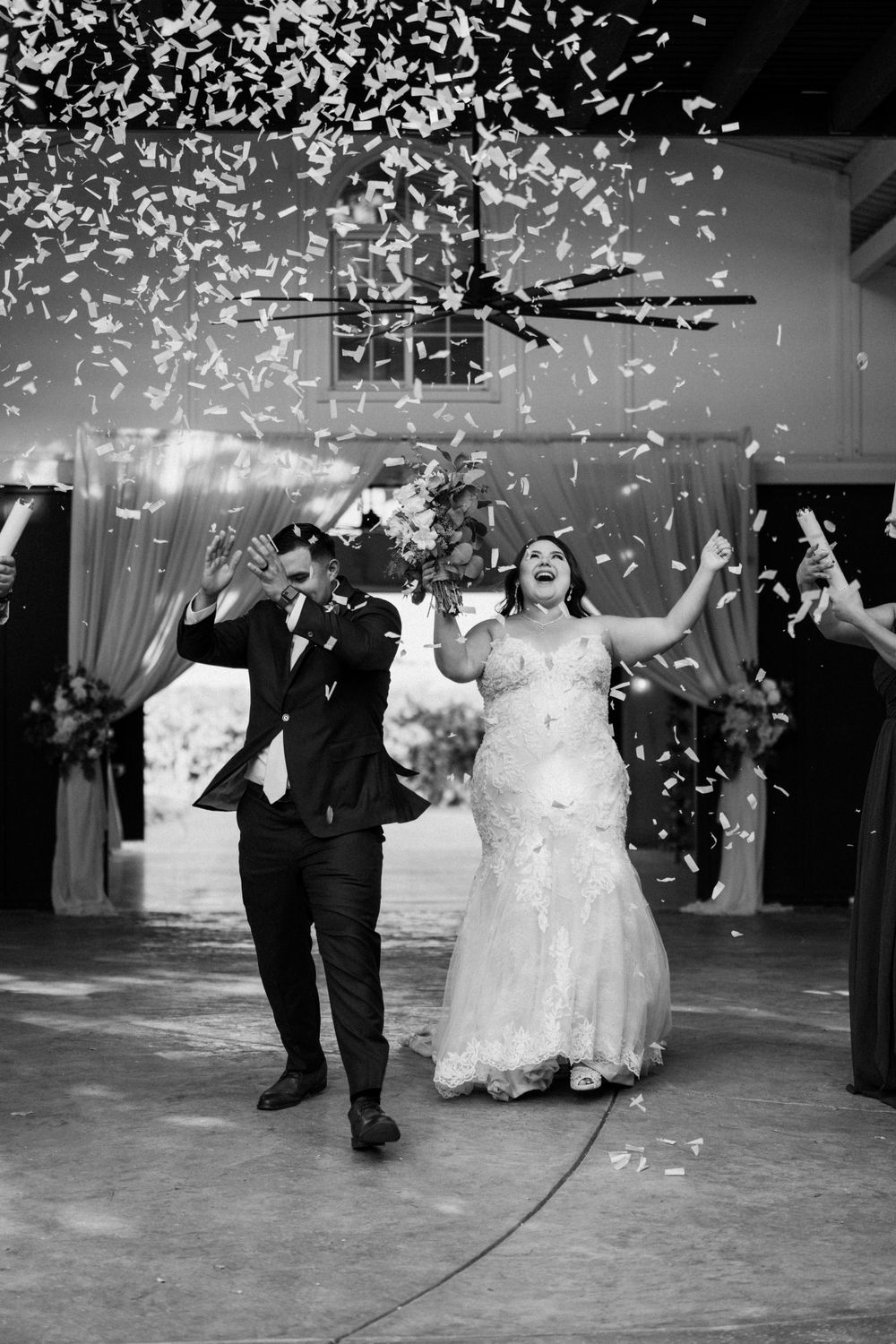 Black and white photo of newlyweds celebrating under shower of confetti at reception.