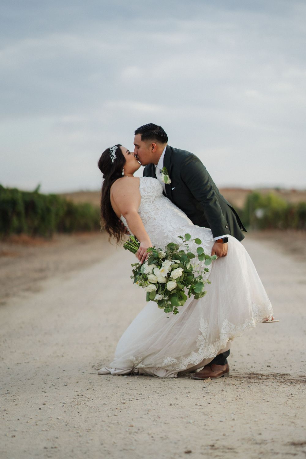 A romantic wedding couple shares a kiss on a dirt road while the bride holds a white and green bouquet.
