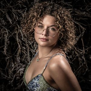 Portrait of curly-haired person in glasses and floral top against dark natural background. Portrait photography.