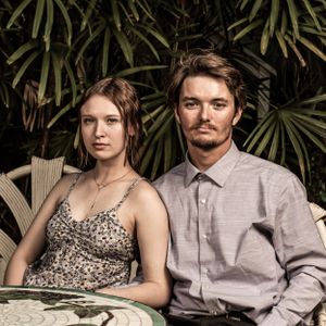 Couple in formal attire poses at outdoor table with tropical foliage background. Family portrait photography.