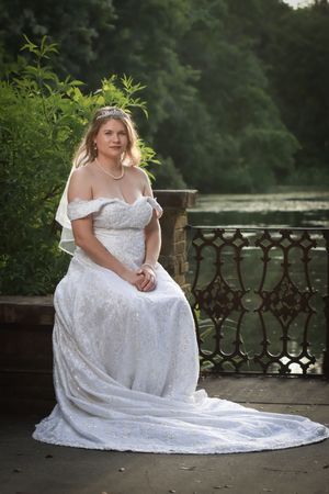 A bride in an off-shoulder lace wedding gown with long train sits on a stone balcony overlooking a serene lake.