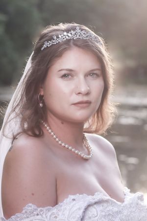 Bride wearing delicate pearl necklace, crystal tiara and white lace dress in dreamy backlit portrait.