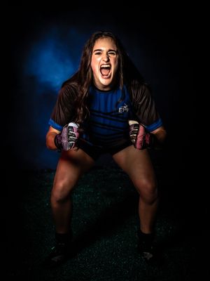 Female rugby player poses with two energy drinks in dramatic lighting against dark blue background.