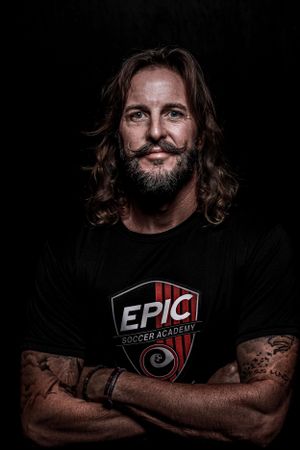 Studio portrait of a bearded person wearing an EPIC Soccer Academy shirt against a dark background. Portrait photography.