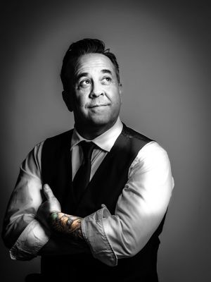 Black and white portrait of a person in business attire with vest and tie looking upward thoughtfully against dark background.