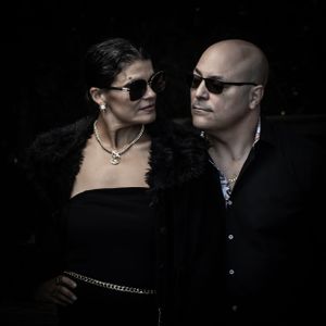 Dramatic portrait of couple in black attire and sunglasses against dark background. Family portrait photography.