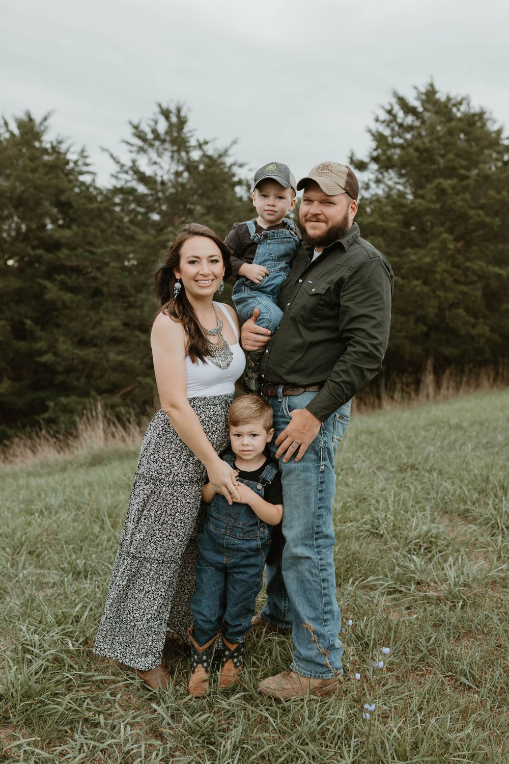 A family of four poses together on a grassy hill with evergreen trees in the background.