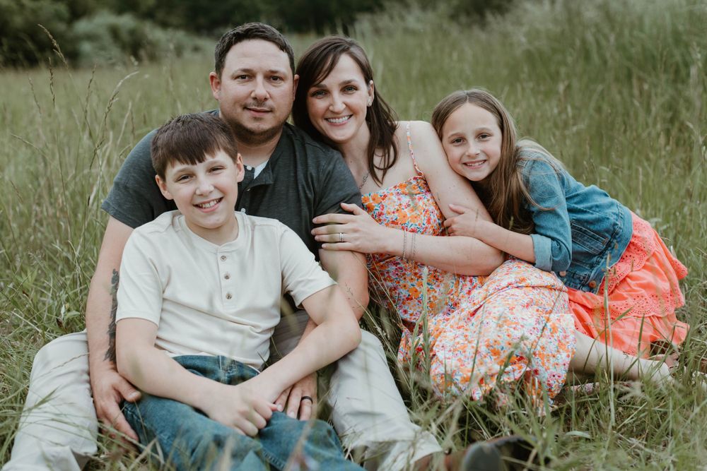 A family of four sitting close together in a grassy field during a casual outdoor photo session.