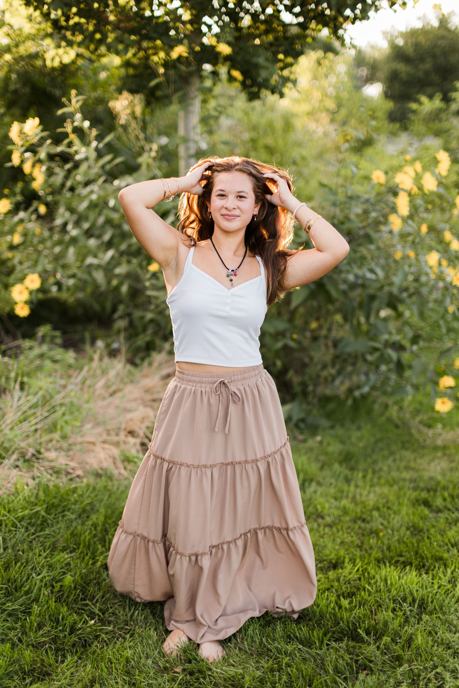 Person in a flowing beige tiered dress posing playfully on grass during golden hour. Twin Cities senior photography.