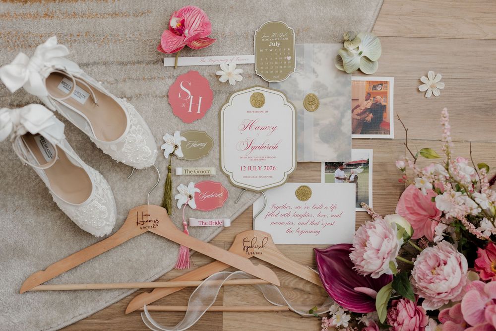 Elegant wedding flat lay with lace bridal shoes, pink floral invitations, wooden hangers, and fresh pink peonies on neutral background.