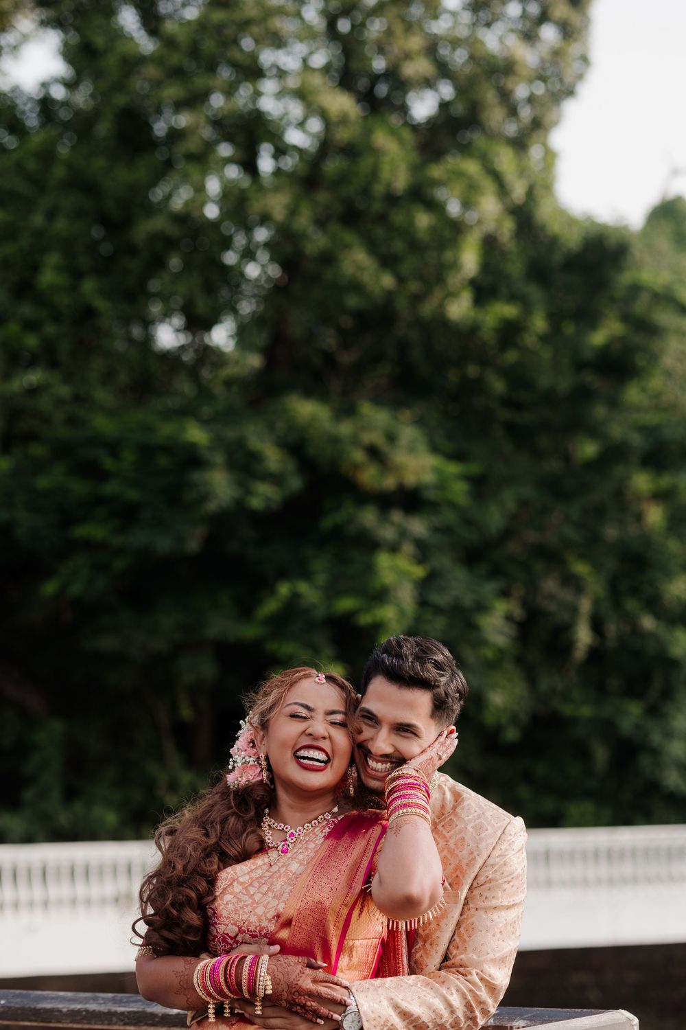A joyful Indian bride in red saree and groom in gold sherwani laughing together outdoors with lush green trees behind them.