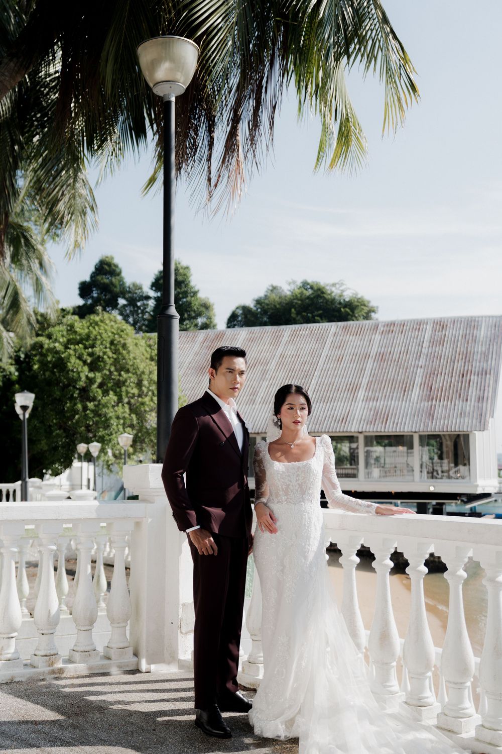 Couple in wedding attire pose on white balustrade, groom in burgundy suit, bride in lace gown, tropical setting.