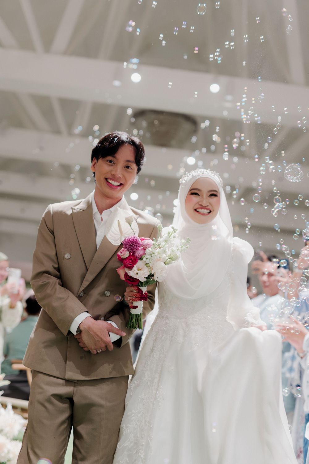Smiling newlywed couple walks down aisle surrounded by bubbles, groom in tan suit, bride in white hijab holding floral bouquet.