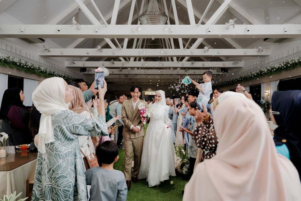 Muslim wedding couple walks through cheering guests in a decorated indoor venue with white beamed ceiling.