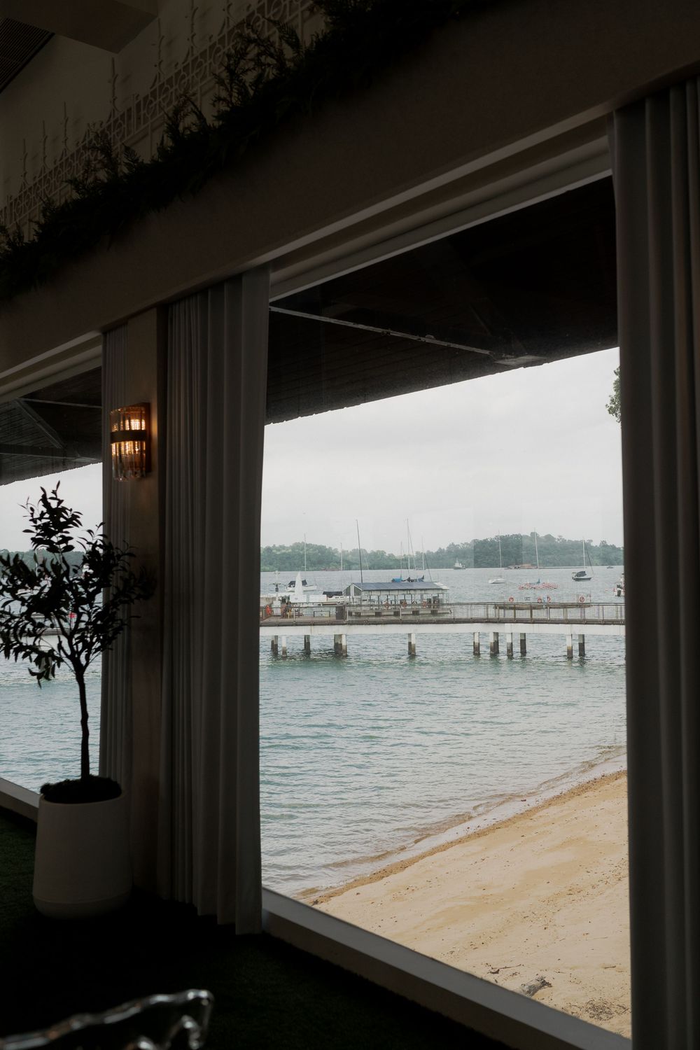 View from an elegant waterfront venue through large windows overlooking a sandy beach, pier, and calm bay on an overcast day.