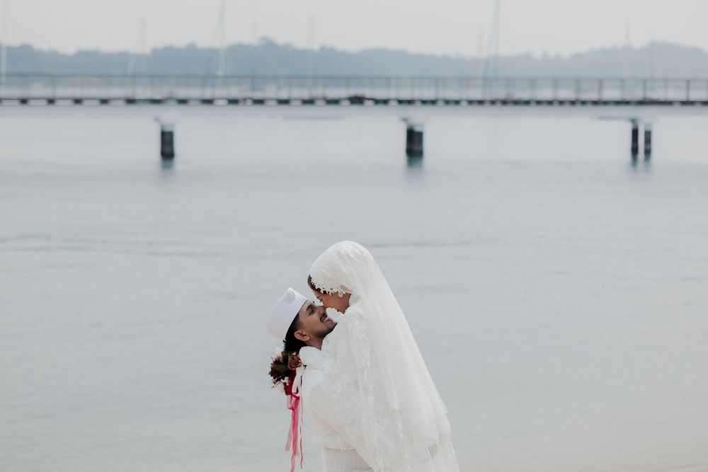 Bride in white hijab and wedding dress poses gracefully by a calm waterfront with a pier in the background.