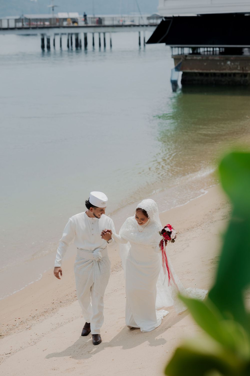 Muslim couple in white wedding attire walking hand in hand on a tropical beach near a pier.