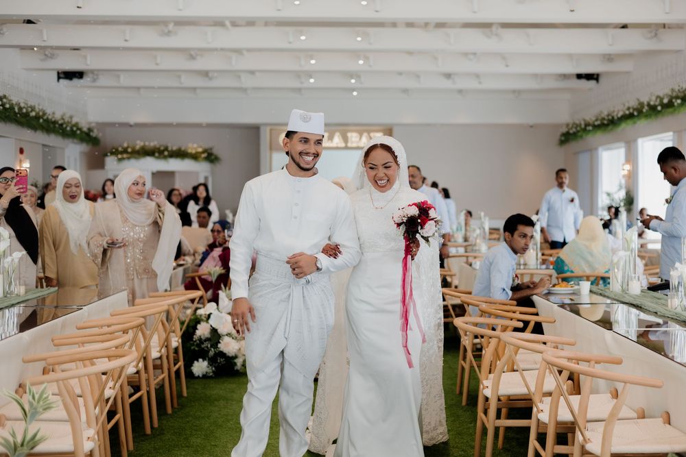 Muslim couple in white wedding attire smiling at their elegant reception with guests seated at wooden chairs.