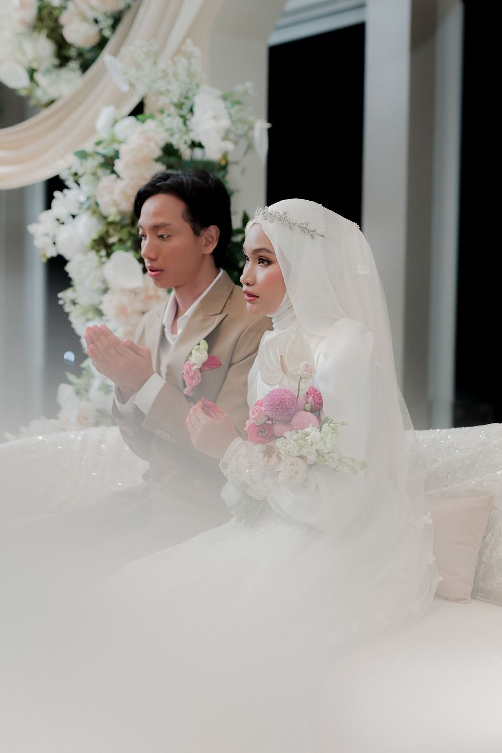 A bride in hijab and groom in beige suit pray together at elegant white floral wedding ceremony.