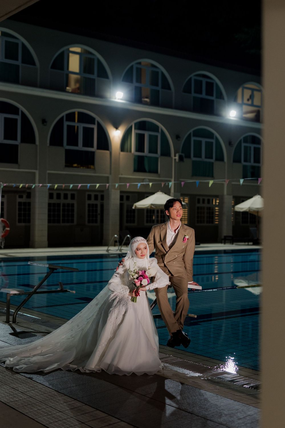 Bride in white hijab gown and groom in tan suit pose by illuminated pool at night wedding venue.