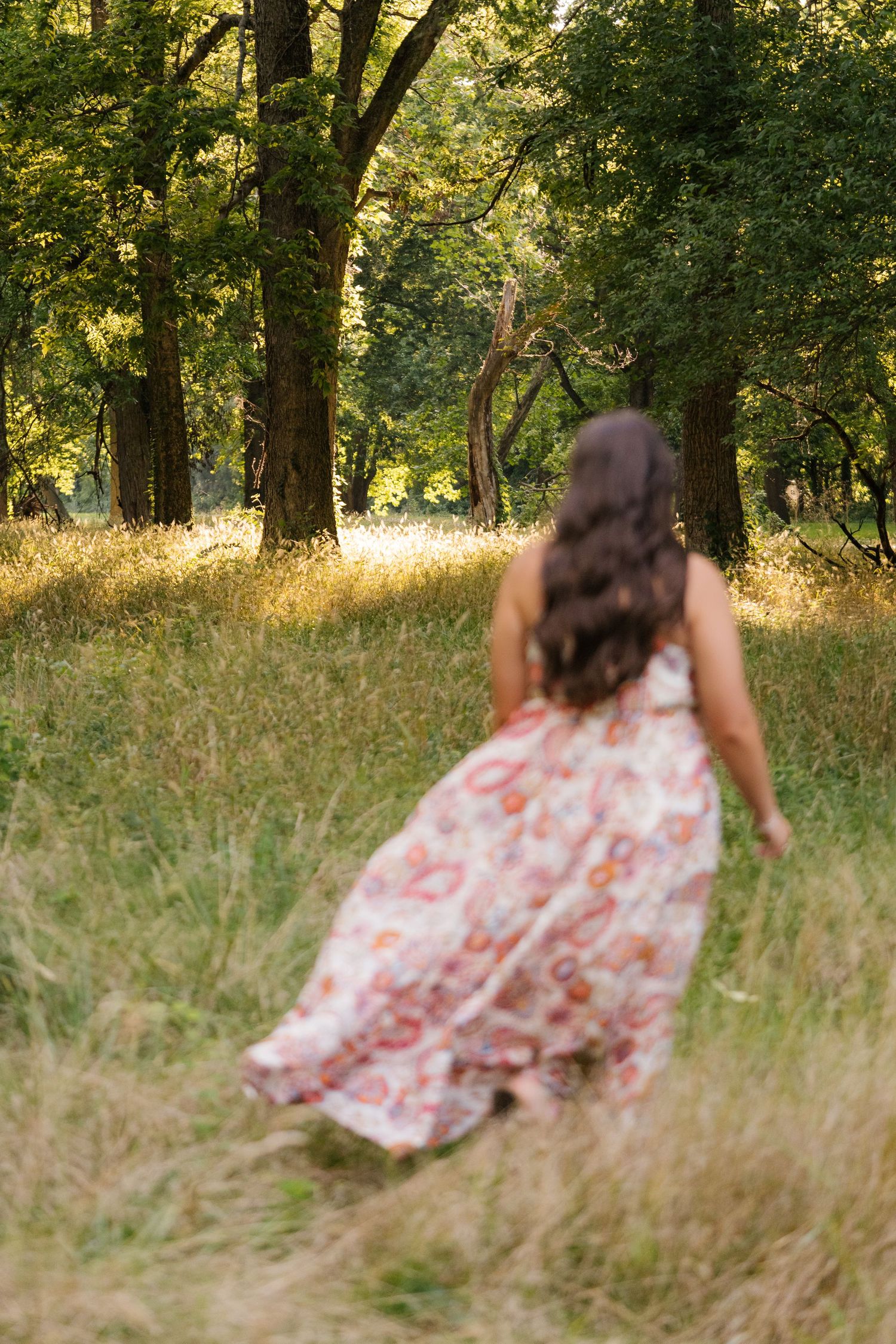 Woman in floral maxi dress walking through a sunlit forest clearing with tall grass and green trees.