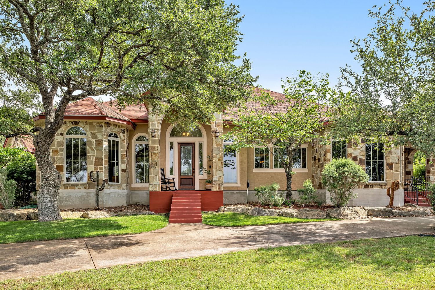 Charming stone and stucco home with mature trees, arched windows, red steps, and lush landscaped front yard.