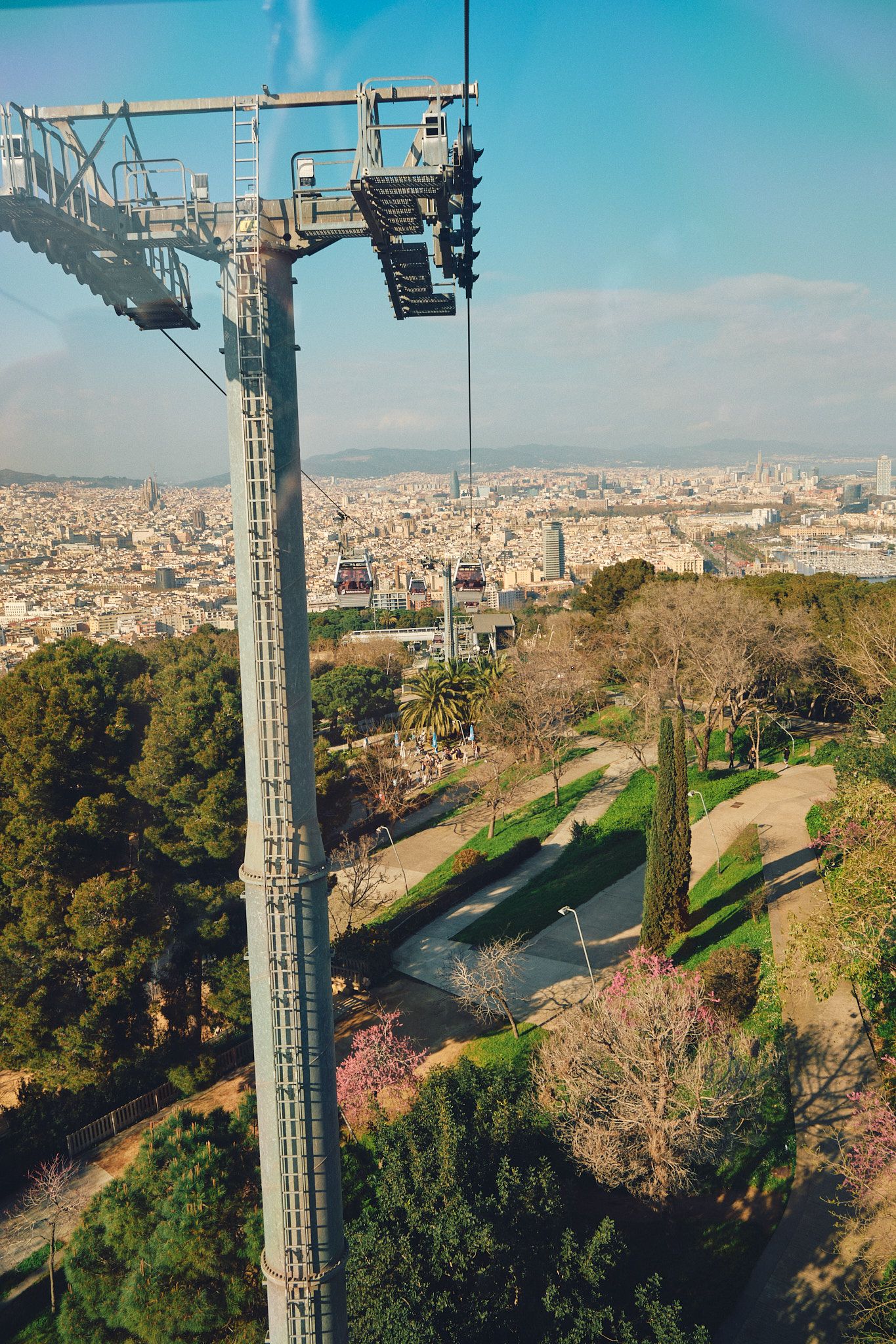 Aerial cable car tower overlooking Barcelona cityscape with lush green park and distant Sagrada Familia visible.