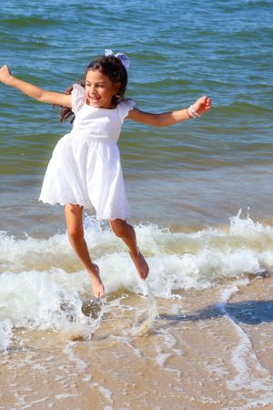 Happy girl in white dress jumps over splashing ocean waves on a sunny beach.