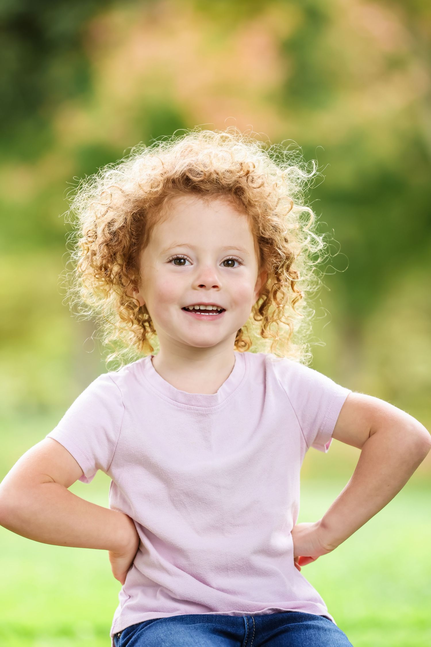 Cheerful young girl with curly blonde hair posing outdoors in a pink t-shirt with hands on hips.