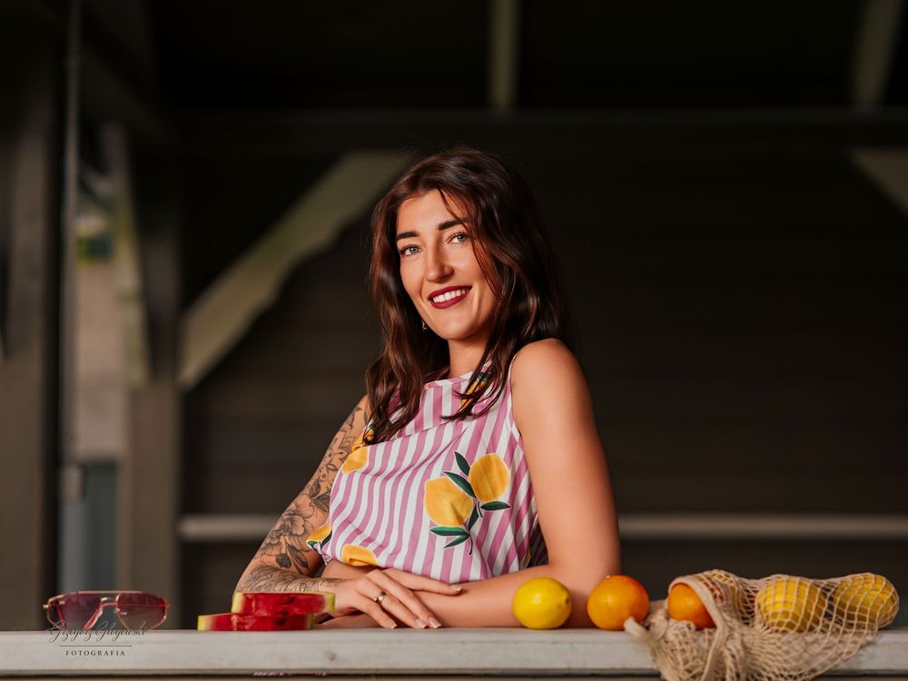 Smiling woman in striped lemon-print dress leaning on railing with fresh fruits nearby.