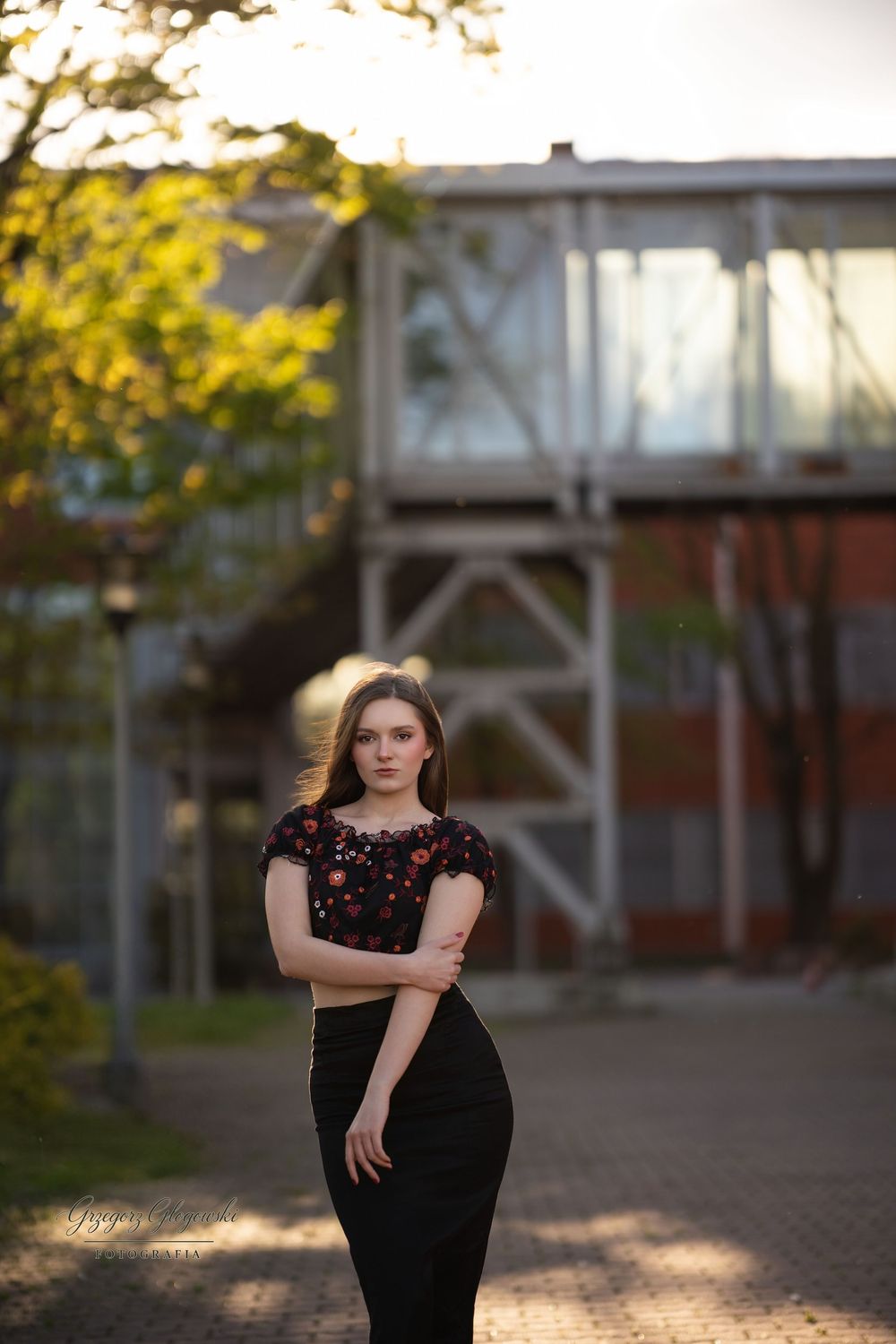 Young brunette woman in black floral crop top and skirt posing outdoors near building.