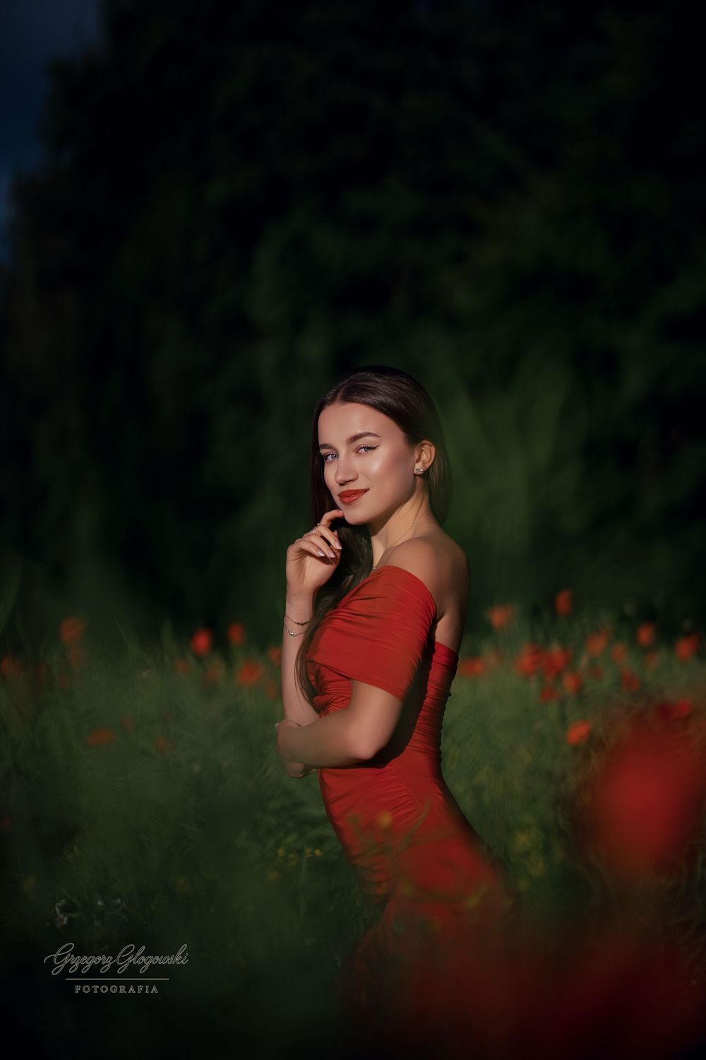 Young woman in red dress stands among red poppies in moody evening light.