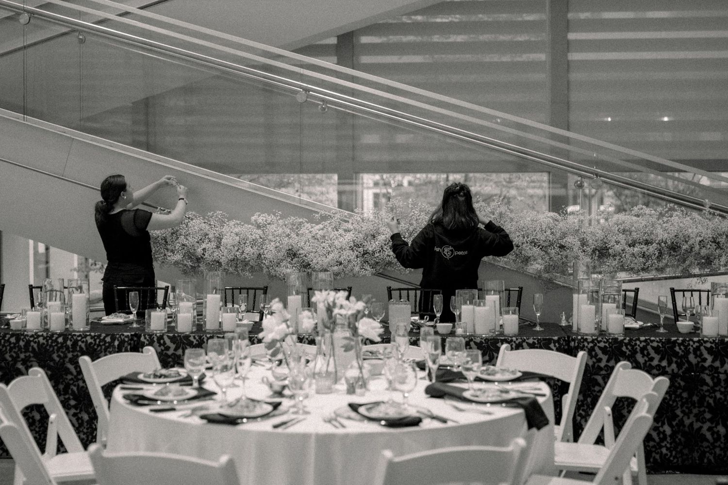 Black and white photo of an elegant wedding reception with round tables, floral centerpieces, and guests mingling in a glass venue.