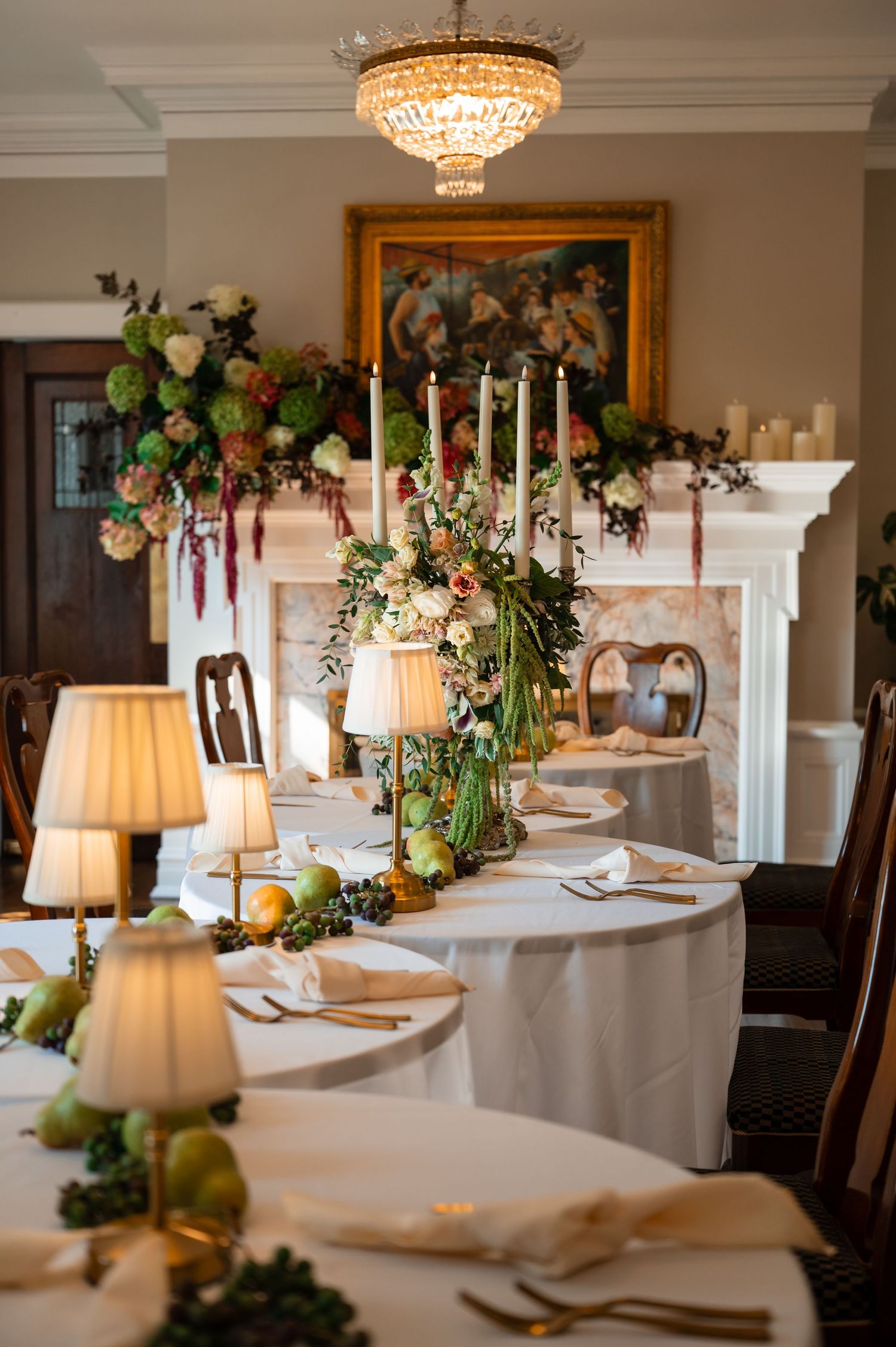 Elegant wedding reception table with floral centerpieces, candles, and fruit accents in a classic dining room with chandelier.