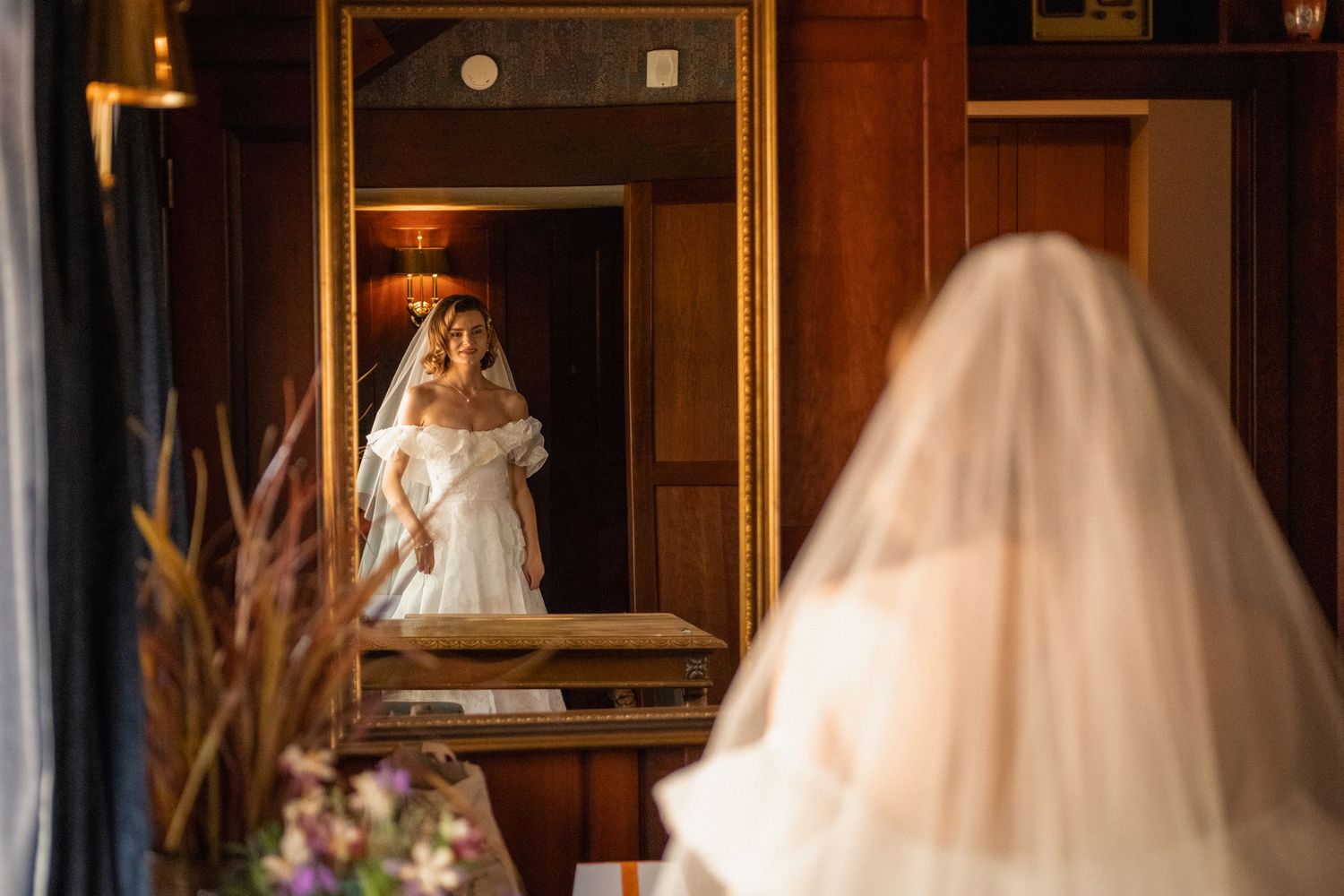 Bride in white gown and veil admires her reflection in an ornate gold-framed mirror in an elegant wood-paneled room.