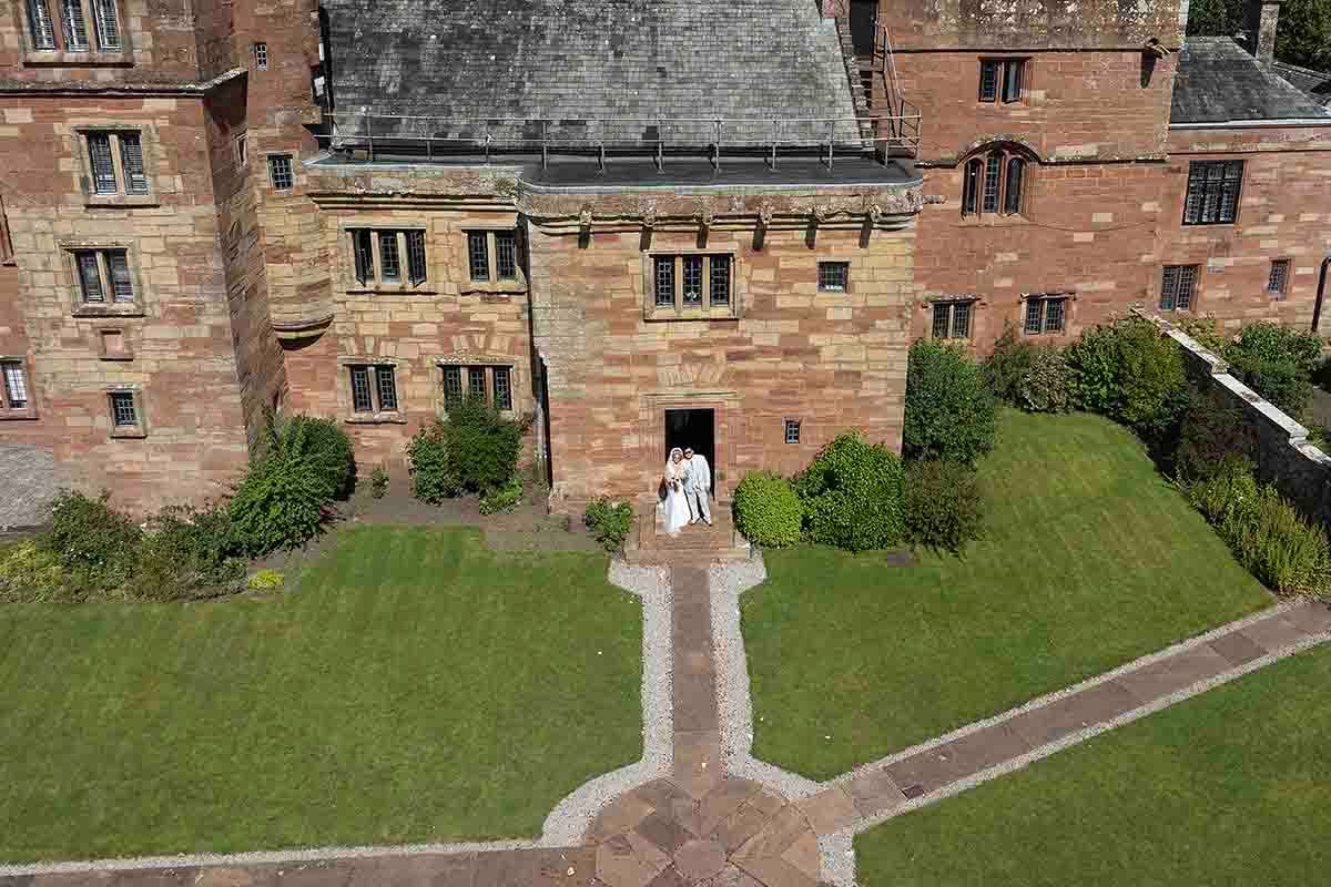 Aerial view of a historic red sandstone castle with manicured lawns and two figures standing at the entrance.