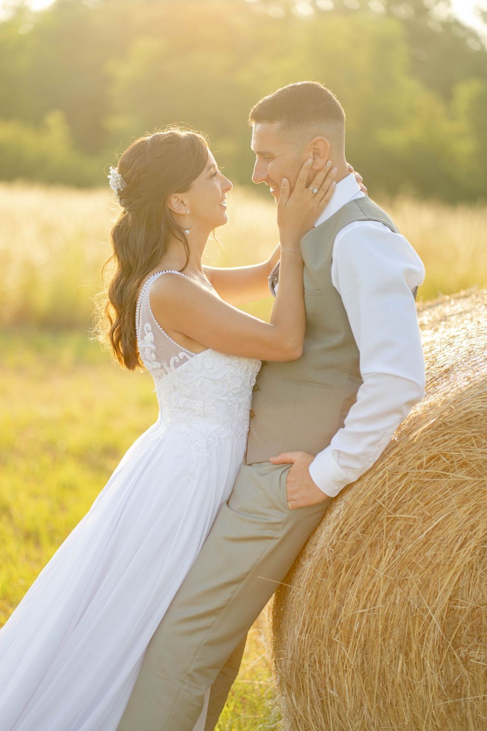 Bride and groom share a romantic moment by a hay bale in a sunlit field, bride in lace gown touching groom's face.