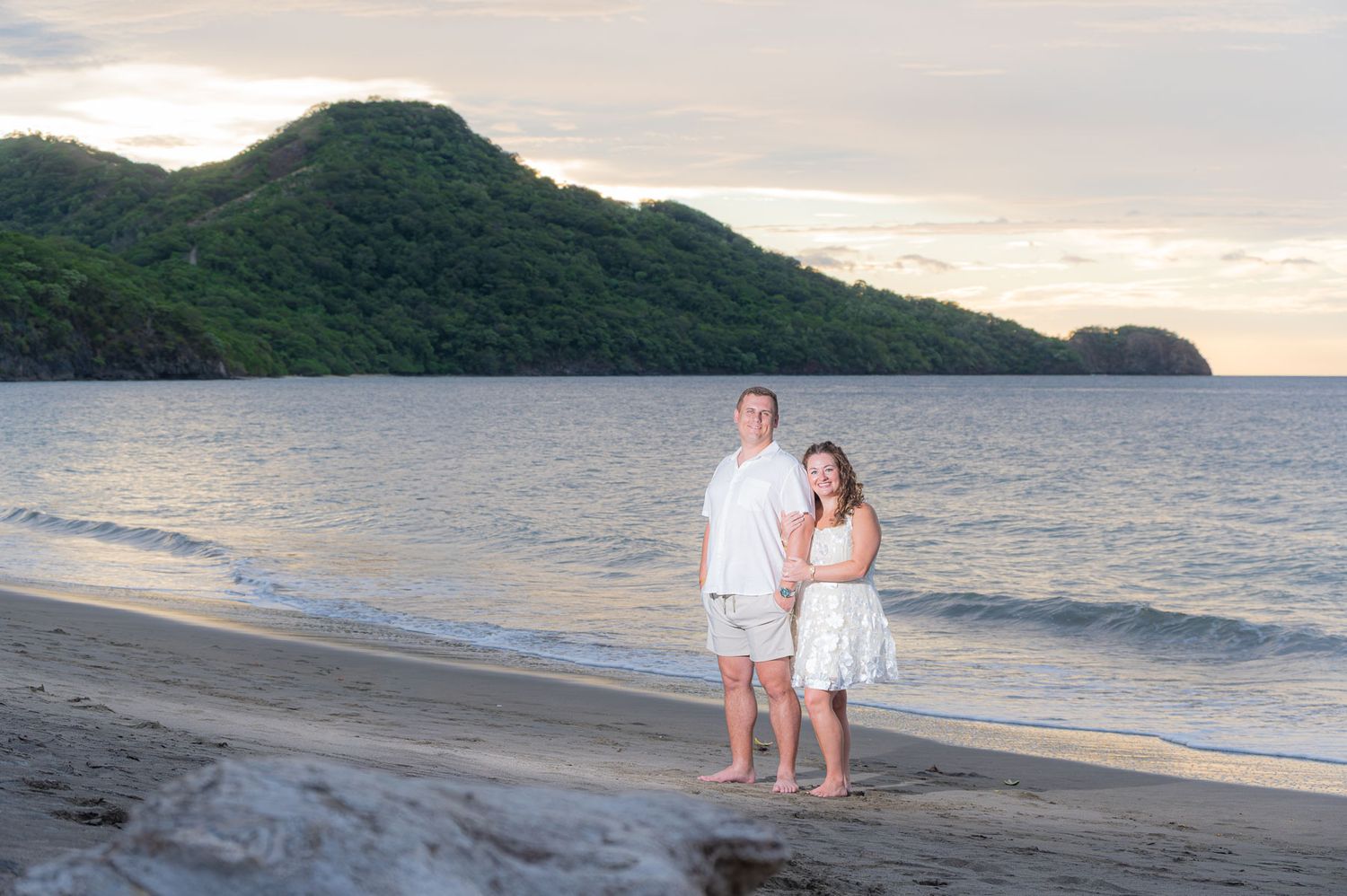 Couple in white outfits standing on a tropical beach at sunset with lush green mountains in the background.