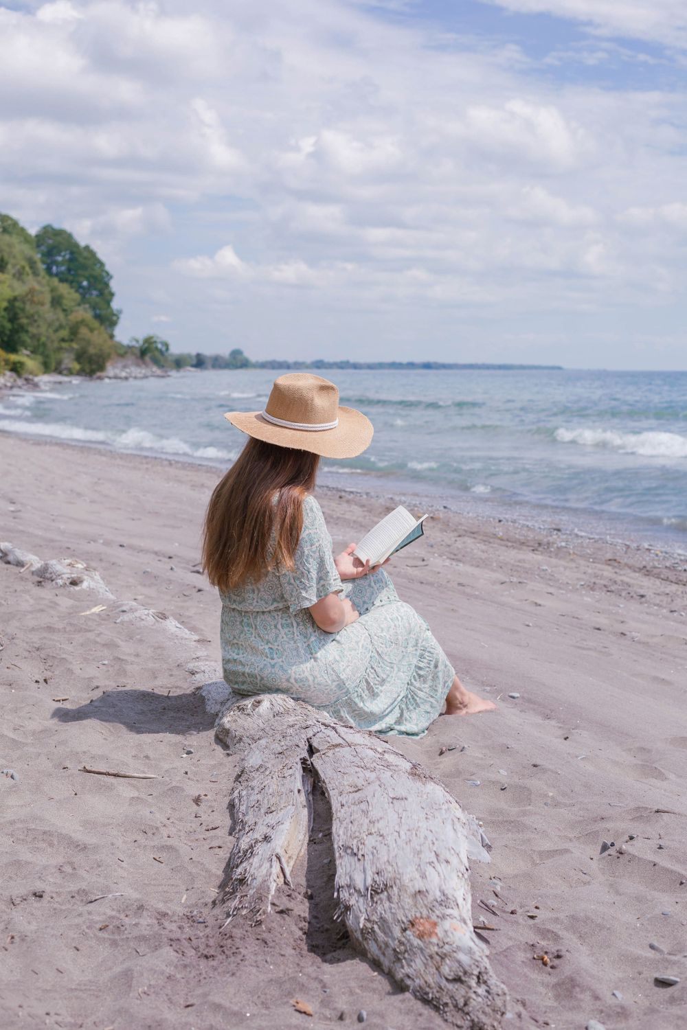 A person in a straw hat reading on a driftwood log at a scenic beach.