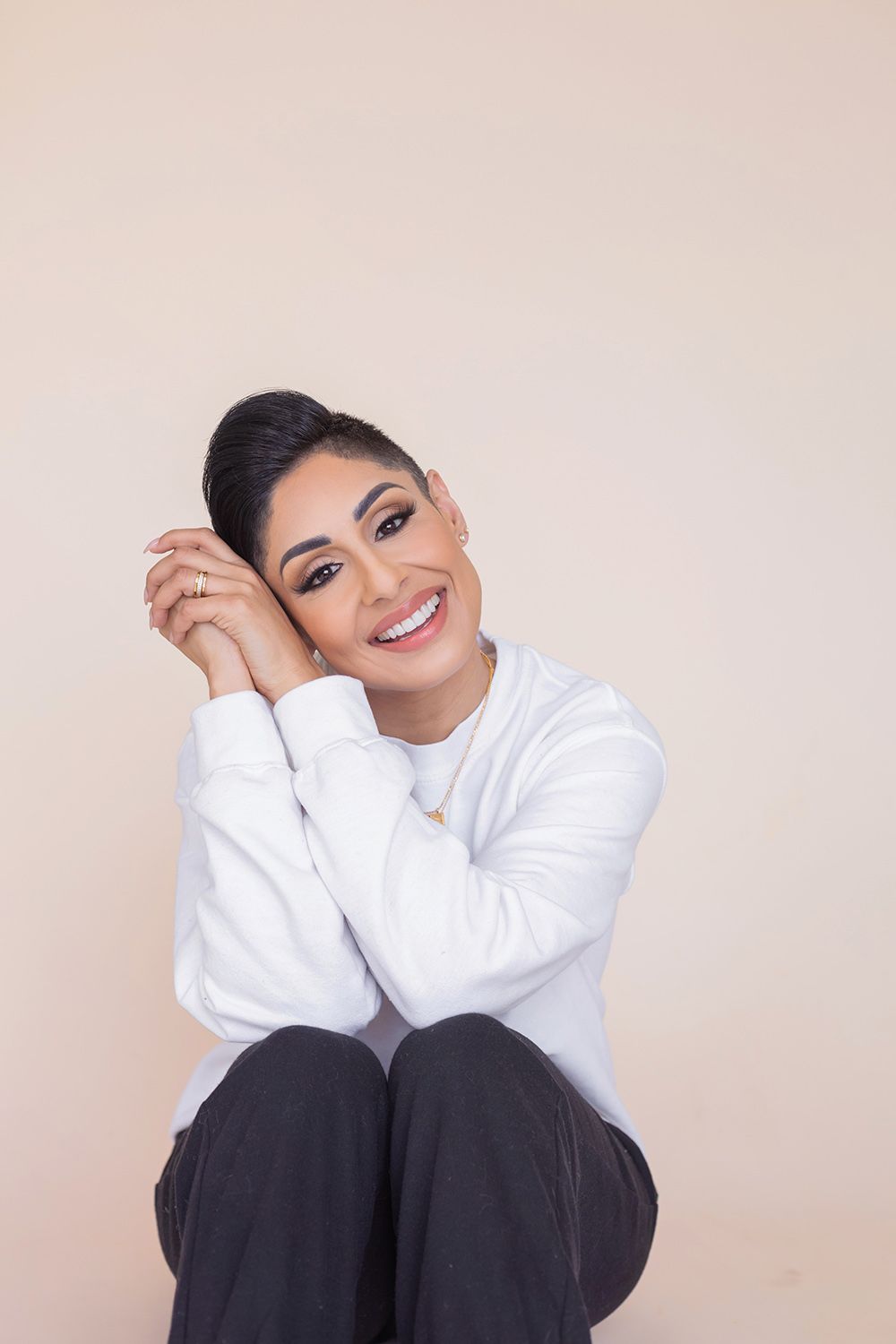 A cheerful person in a white sweater and dark pants sits casually while leaning on their hands against a beige background.