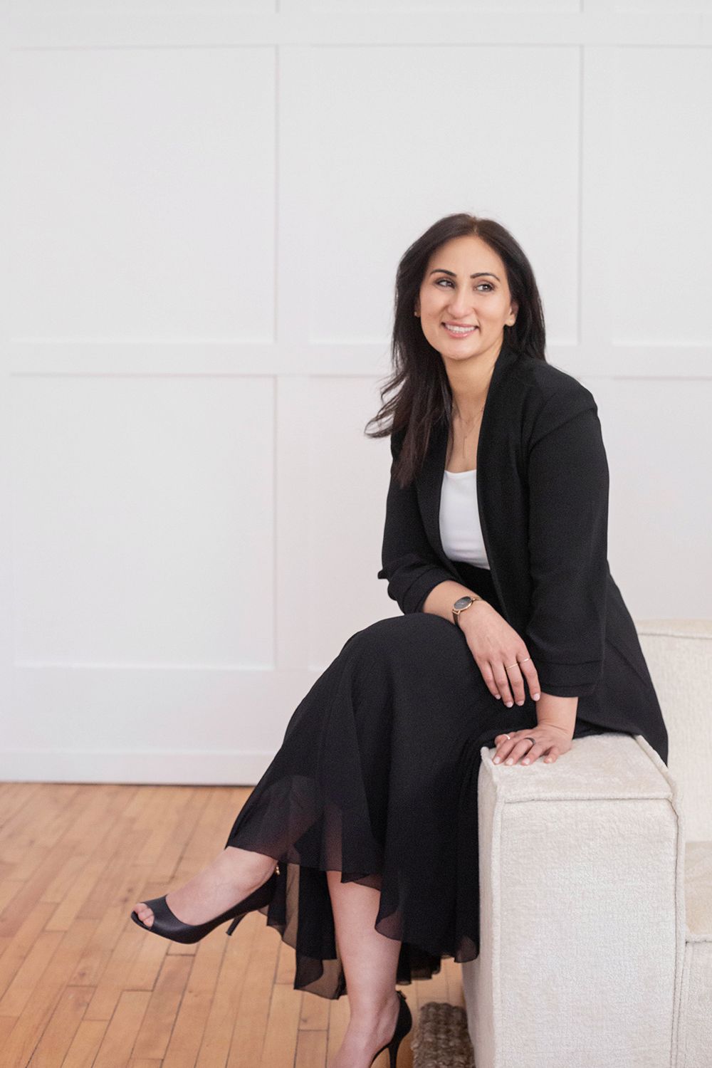 Professional in black blazer and dress sitting elegantly on white furniture.