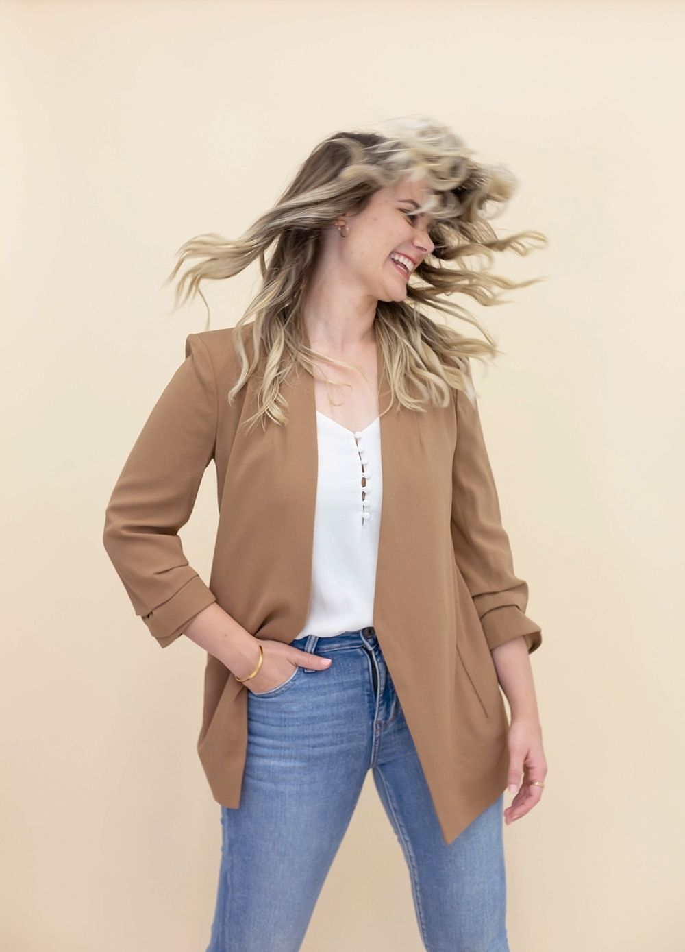 Woman with flowing blonde hair wearing tan blazer and jeans in dynamic motion pose.