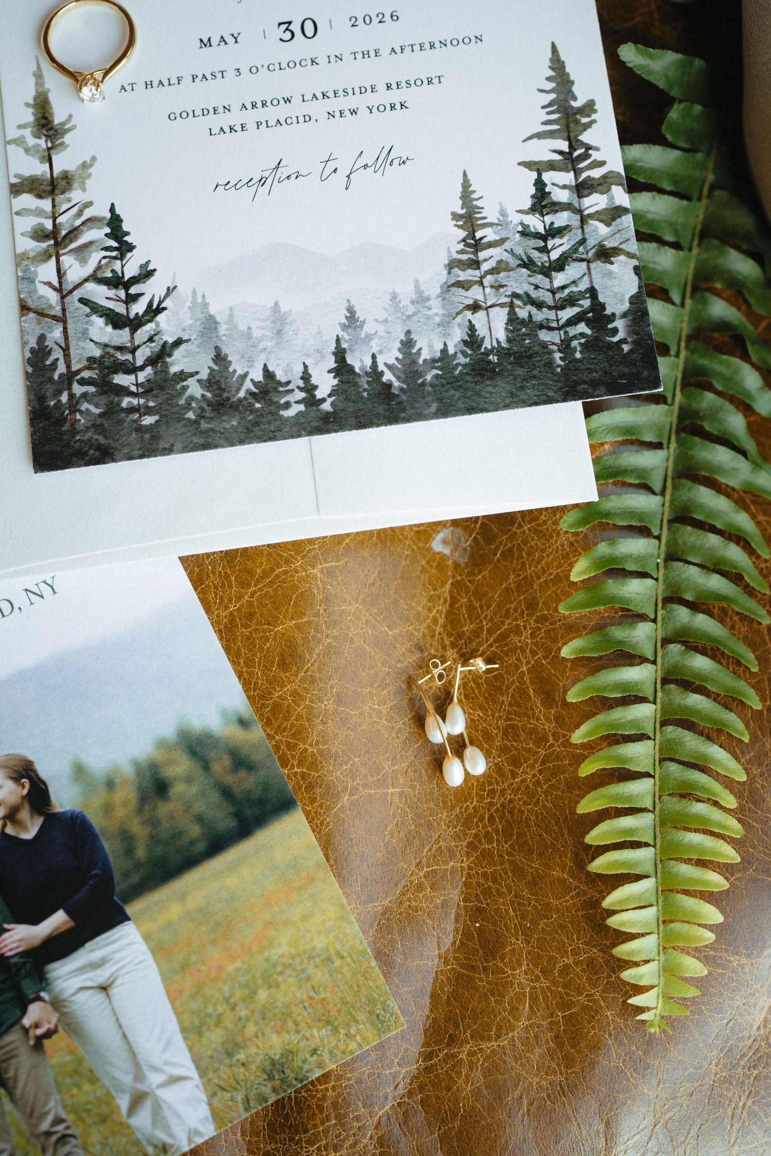 Wedding invitation suite featuring pine tree watercolor design, gold earrings, and fern leaf for Lake Placid NY ceremony.