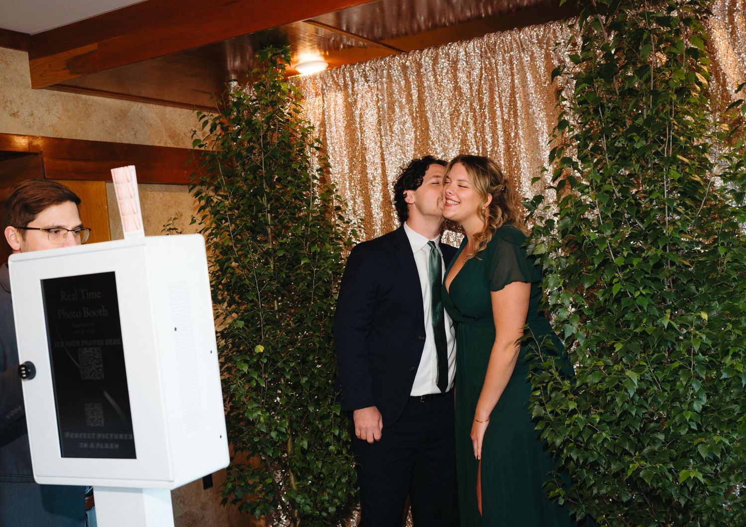 Couple sharing a kiss near a photo booth at an elegant wedding reception with greenery backdrop.