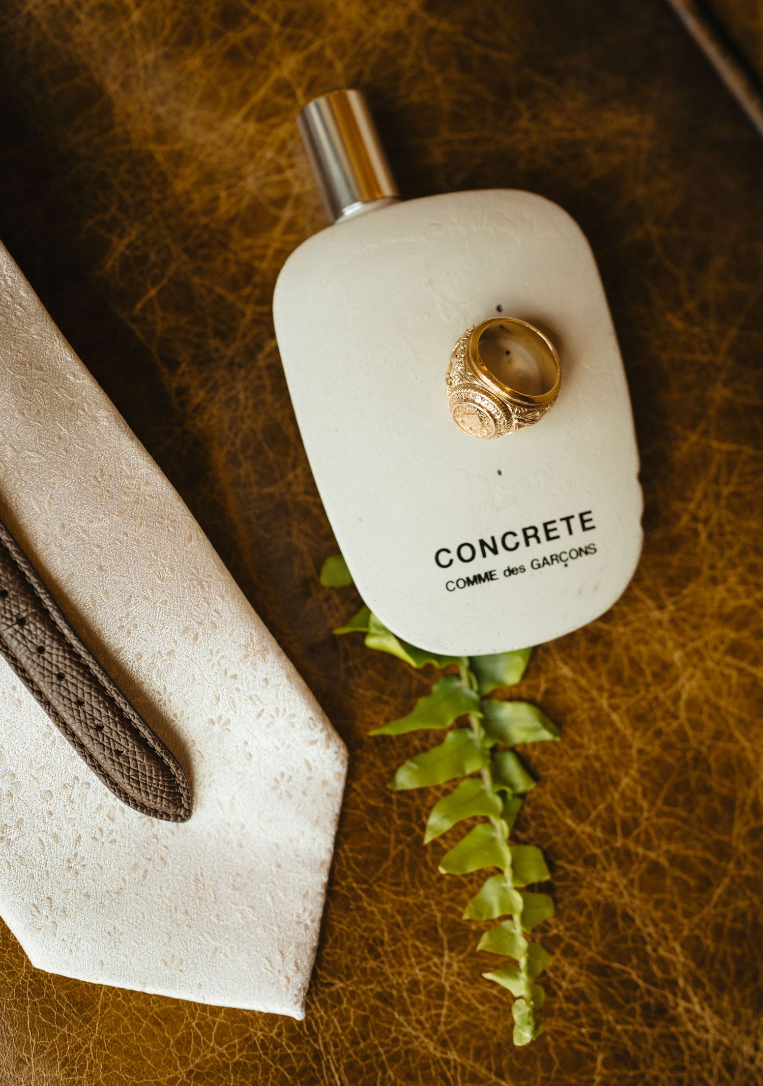Concrete Comme des Garcons perfume bottle with gold ring and green plant on brown leather surface.