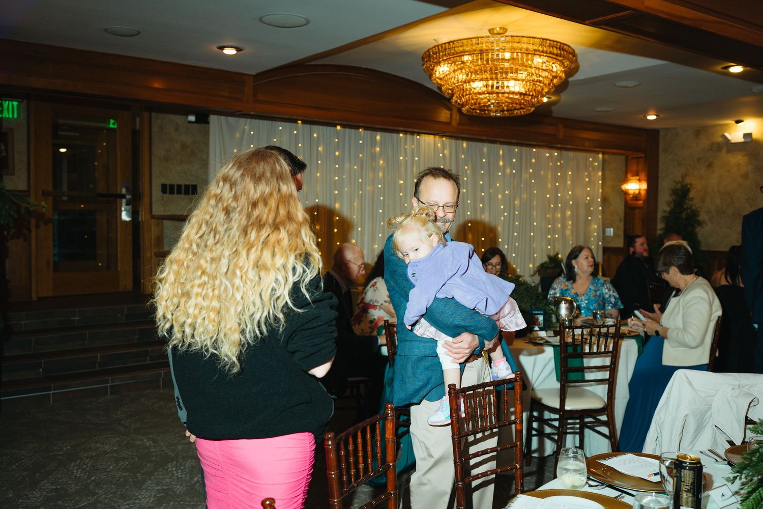 Man holding a baby greets a curly-haired woman at a formal wedding reception with chandelier lighting.